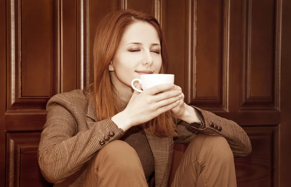 Stijl roodharige meisje drinken koffie in de buurt van houten deuren. — Stockfoto