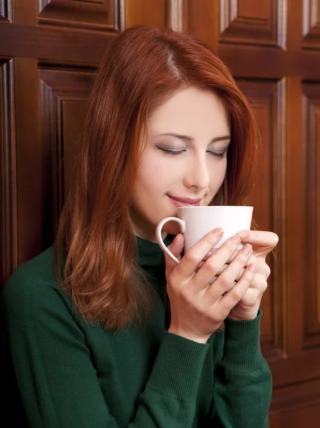 Rödhårig tjej dricker kaffe nära trä dörrar. — Stockfoto