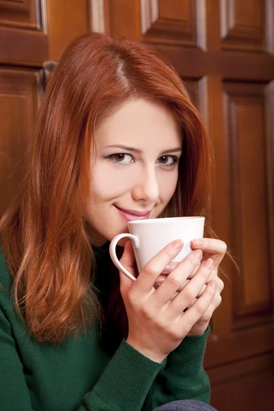 木製のドアの近くのコーヒーを飲むスタイルの赤毛の女の子. — ストック写真