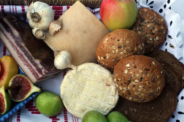 Bodegón rústico de pan, queso, tocino y fruta en una canasta de mimbre —  Fotos de Stock