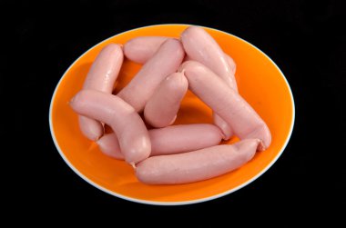 Raw sausages on orange plate against black background clipart