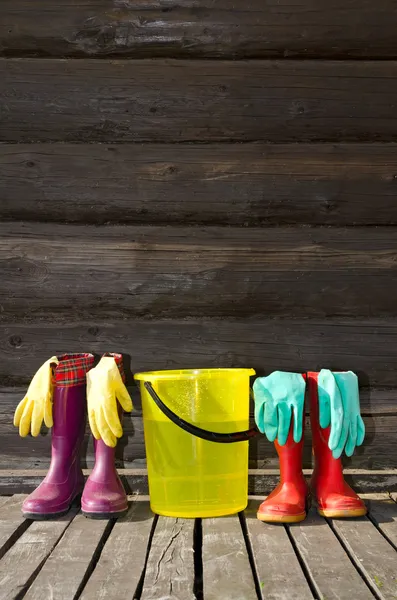 Twee paren van rubber laarzen, emmer en rubber handschoenen op zonnige veranda — Stockfoto