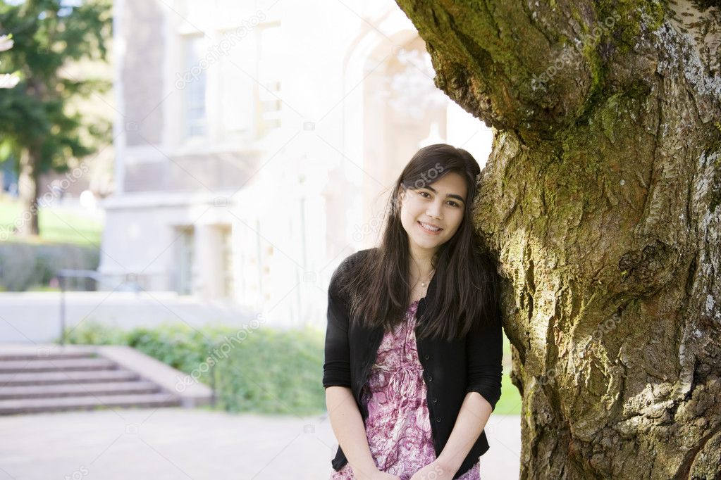 Teen girl leaning against trunk of large tree, smiling