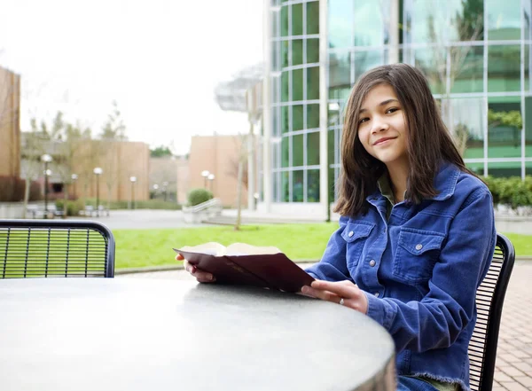 Doze anos de idade menina biracial sentado ao ar livre lendo um livro — Fotografia de Stock
