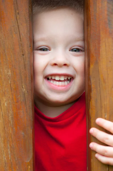 Schattige kleine jongen gluren uit door houten balken glimlachen — Stockfoto