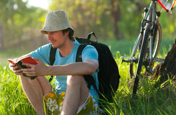 Junger Reisender mit Fahrrad liest das Buch, während er sich an einem sonnigen Frühlingstag im Wald ausruht — Stockfoto