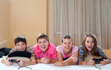 Happy teenage boys and girls lying on bed and playing computer games clipart