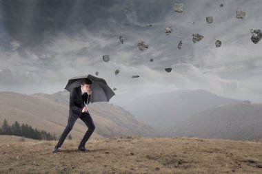 Businessman sheltering under an umbrella from rocks falling to the ground clipart