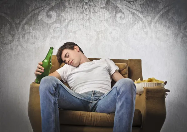 Mladý muž spí na křeslo zatímco drží láhev piva a misku lupínků — Stock fotografie