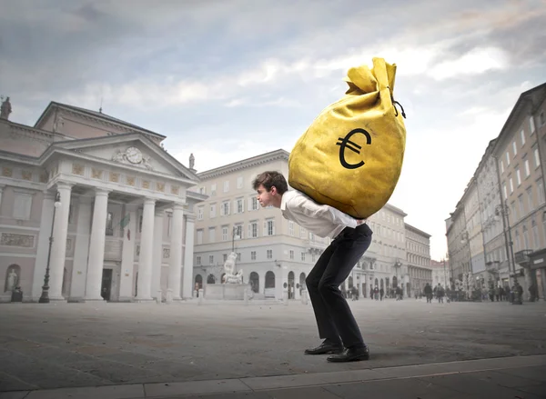 Jovem empresário carregando um saco de dinheiro em seus ombros em uma praça da cidade — Fotografia de Stock