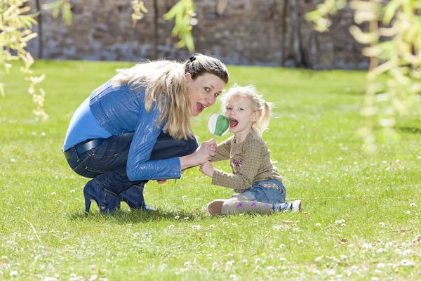 Mother and her daughter with a lollipop sitting on grass — Stock Photo, Image