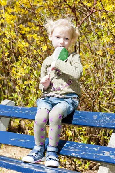 在春天坐在长凳上一根棒棒糖的小女孩 — 图库照片
