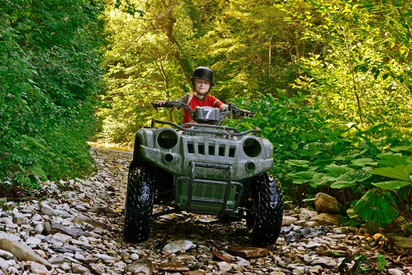 A little boy wearing a helmet riding a quad bike on the shore of a mountain river. — Stock Photo, Image
