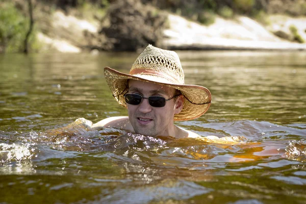 A man in a hat and sunglasses swimming in a river. — Stock Photo, Image