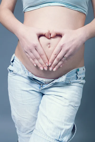 Beautiful pregnant white woman wearinglight blue top and jeans. Hands on belly, fingers in heart shape over navel. Pregnancy collection. — Stock Photo, Image