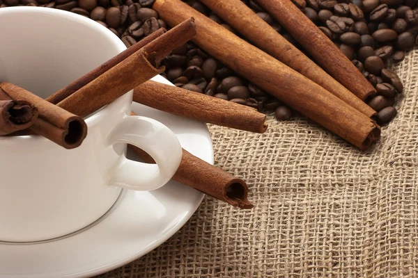White coffee mug, beans and cinnamon sticks on sacking — Stock Photo, Image