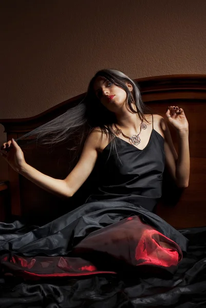 Piękna młoda kobieta w czarno -czerwona sukienka, siedząc na łóżku — Zdjęcie stockowe