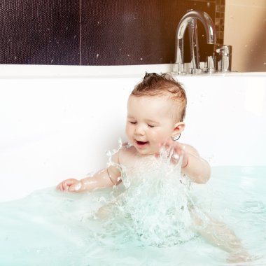 Cute baby having fun, splashing water and laughing while taking a bath in a modern bathroom interior clipart