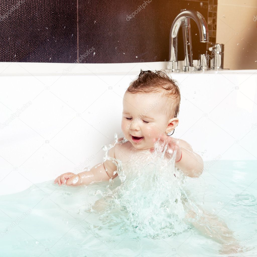 Cute baby having fun, splashing water and laughing while taking a bath in a modern bathroom interior
