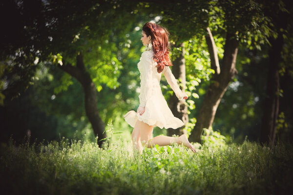 Redhead woman illuminated by sunlight in elegant dress barefoot running through the woods