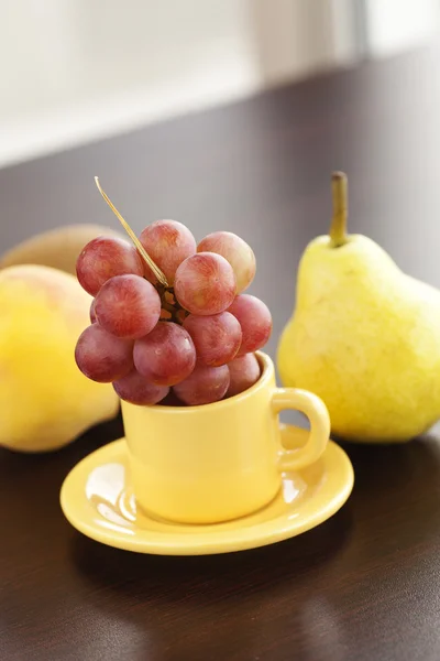 Pesca, pera, uva in una tazza e piattino su un tavolo di legno — Foto Stock
