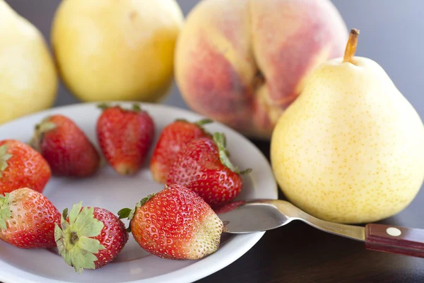 Jordgubbar i tallrik, sked, päron och persikor på ett träbord — Stockfoto