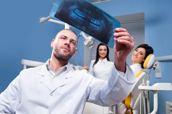 Dentista com raio-x e paciente sorridente com assistente em segundo plano — Fotografia de Stock