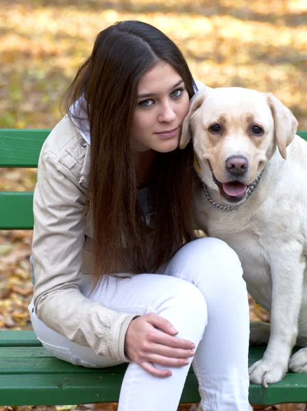 Hermosa chica y su perro sentado en el banco en el parque de otoño — Foto de Stock