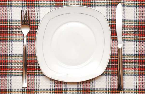 Белая пустая тарелка с вилкой и ножом на клетчатой скатерти — стоковое фото