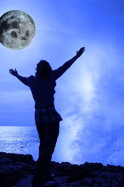 Une femme avec les mains levées face à une vague et la pleine lune sur la falaise e — Photo
