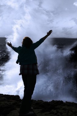 Silhouette of lone woman facing a powerful wave on cliff edge clipart