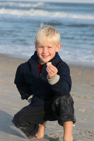 Sonriente chico agrupado en ropa en la playa mostrando Shell — Foto de Stock