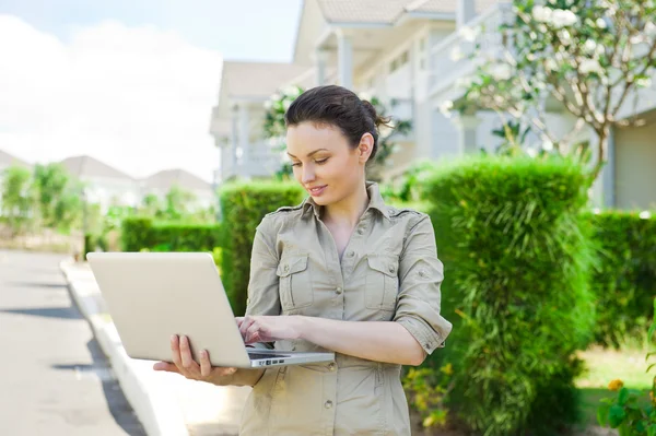 Junge Geschäftsfrau (Immobilienmaklerin) hält einen Laptop in der Hand und präsentiert ein freistehendes Einfamilienhaus — Stockfoto