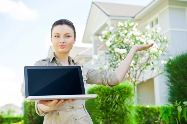 Junge Geschäftsfrau (Immobilienmaklerin) hält einen Laptop in der Hand und präsentiert ein freistehendes Einfamilienhaus — Stockfoto