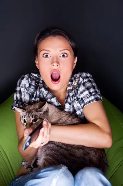 Joven mujer divertida sentada en su bolsa de frijoles y abrazando a su gato mientras ve la televisión. Ella está muy asustada mientras ve una película de terror en un cuarto oscuro — Foto de Stock