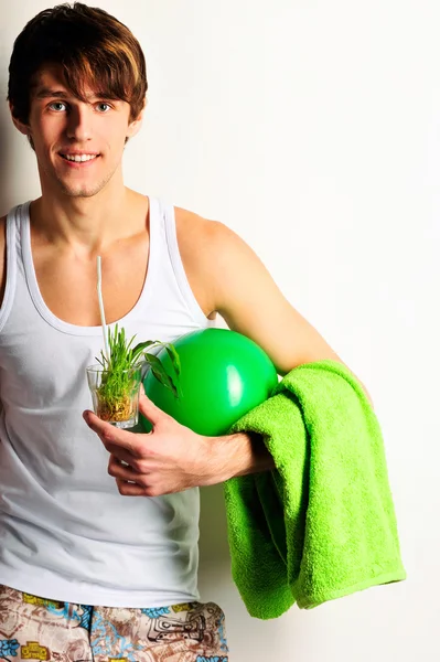 Young healthy european man looking at camera drinking refreshing juice and holding ball and towel. Standing against wall. — Stock Photo, Image