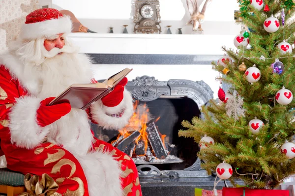 Santa sentado en el árbol de Navidad, cerca de la chimenea y la lectura — Foto de Stock