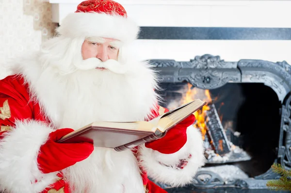 Santa sitting at the Christmas tree, near fireplace and reading Stock Photo