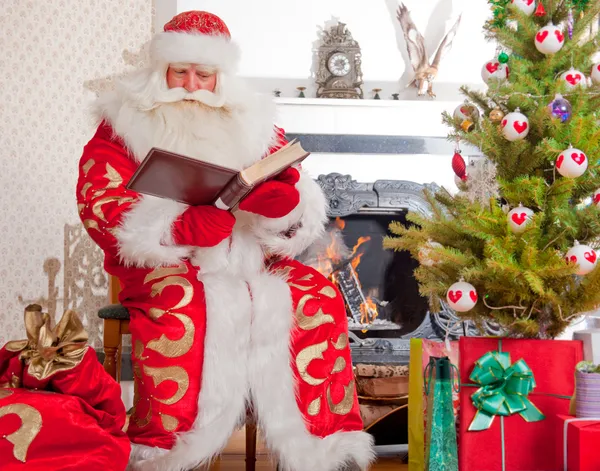 Santa sitting at the Christmas tree, near fireplace and reading Stock Photo
