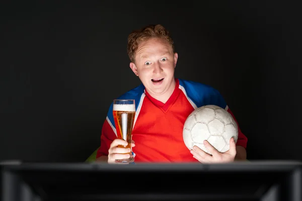 Νεαρός άνδρας κρατά μπάλα ποδοσφαίρου και μπύρα και βλέποντας τηλεόραση translati — Φωτογραφία Αρχείου