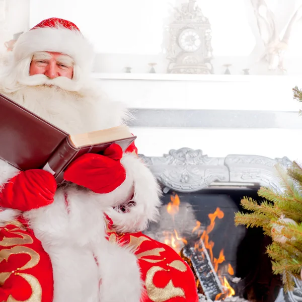 Santa sitting at the Christmas tree, near fireplace and reading — Stock Photo, Image