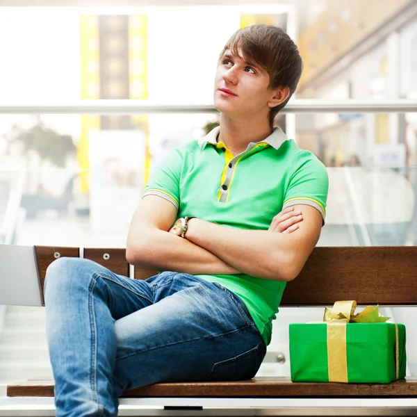 Retrato de hombre joven dentro del centro comercial con caja de regalo sentado —  Fotos de Stock