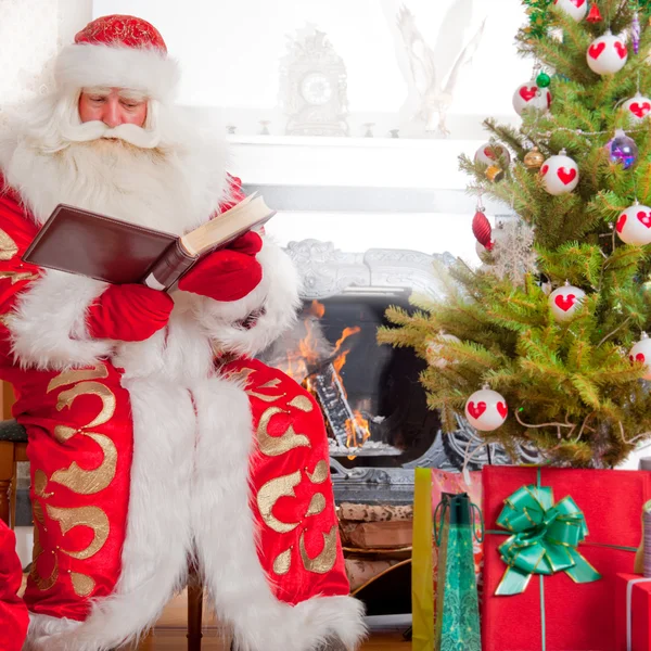 Santa sitting at the Christmas tree, near fireplace and reading Stock Picture