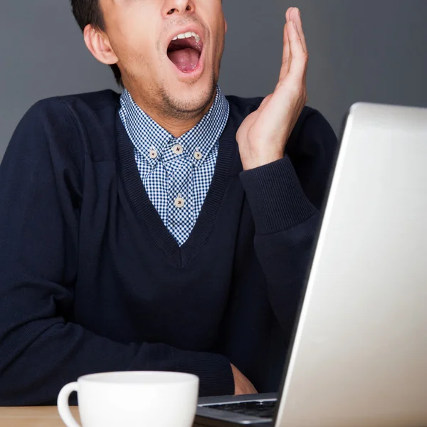 Jeune homme d'affaires bâillant devant un ordinateur portable et une tasse de thé ou — Photo