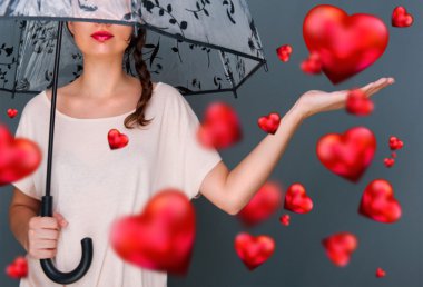Young fashionable woman holding umbrella standing against grey background red hearts are floating around her. Love rain concept clipart