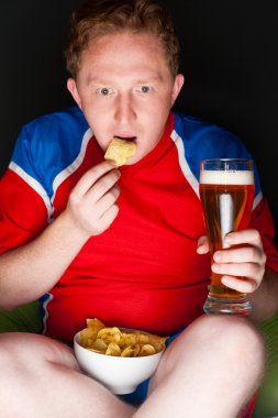 Close-up portrait of young man wearing sportswear fan of football team is watching tv and rooting for his favorite team. Sitting on beanbag alone at night drinking beer and eating chips clipart