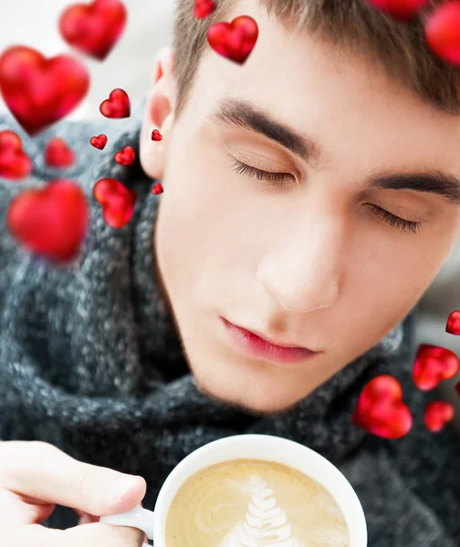 Portrait of a young man drinking coffee while sitting on armchair at home and dreaming about his couple. Red hearts are flying around his head. Valentine concept — Stock Photo, Image