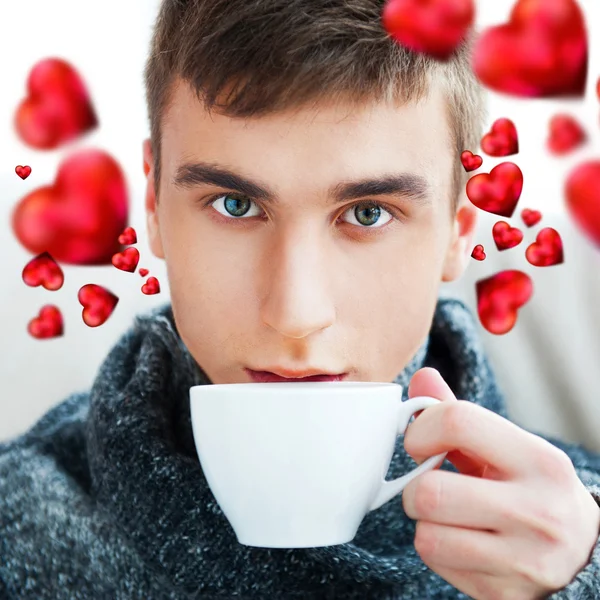 Portrait of a young man drinking coffee while sitting on armchair at home and dreaming about his couple. Red hearts are flying around his head. Valentine concept — Stock Photo, Image