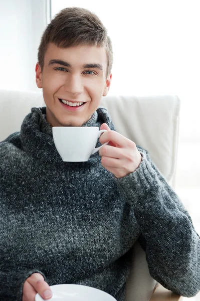 Portrét smějící se mladý muž, pití kávy, zatímco sedí na křesle doma — Stock fotografie