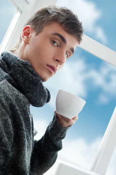 Portrait of a young man drinking coffee and looking at camera, bright sky and clouds outside — Stock Photo, Image